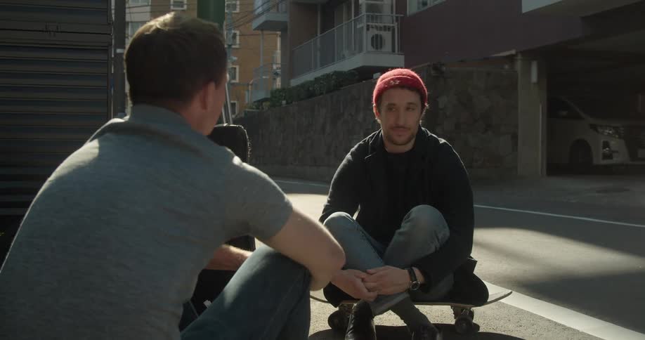 Two white males sit and talk in a neighborhood as a bicyclist rides by in the background