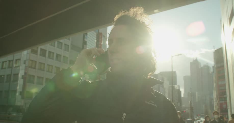 A man stands in a city talking on a phone as pedestrians pass by him