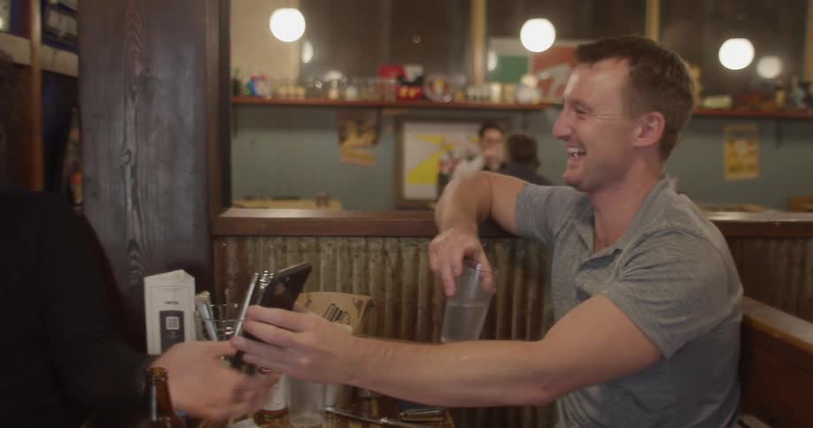 Two men sit at a table in a restaurant and look at a cell phone and laugh