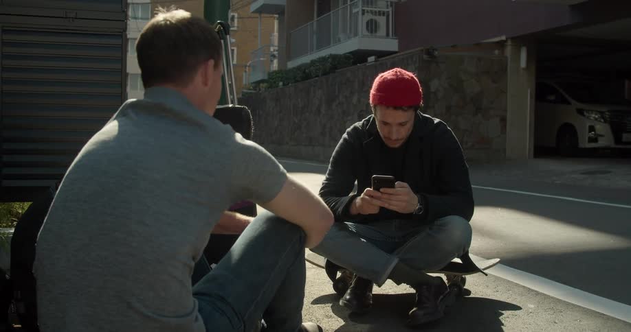 Two friends sit and talk on a neighborhood street as one of the men uses his cell phone