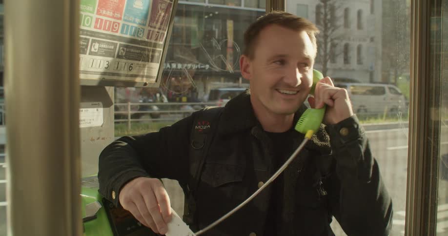 A man smiles as he talks on a payphone while cars and trucks drive past on a sunny day in the city