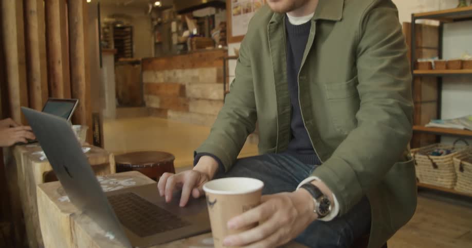 A man sits at a small cafe counter drinking a cup of coffee and working on a laptop