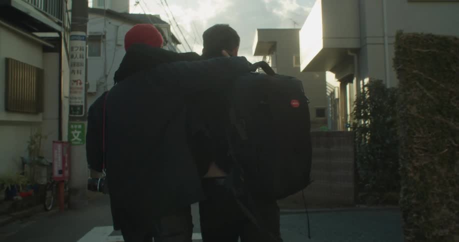 Two white males embrace as they walk down a narrow street in a neighborhood