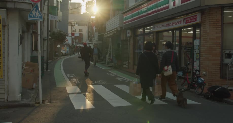 A man skateboards down a narrow road passing people carrying bags and shopping