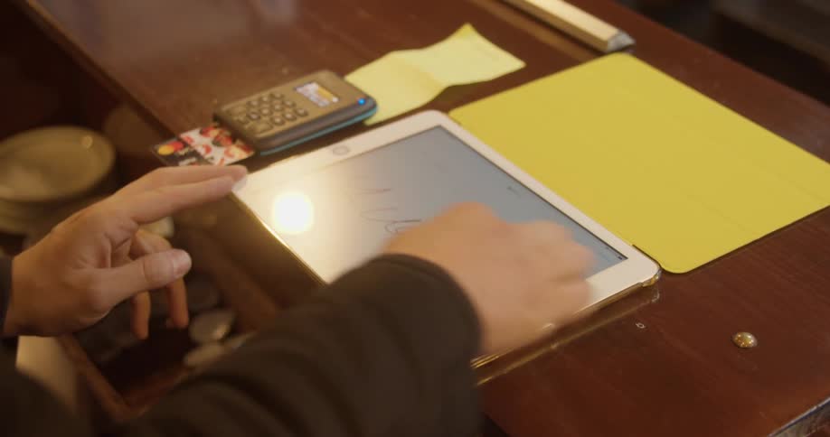 A white man sits at a desk and signs a name on a tablet