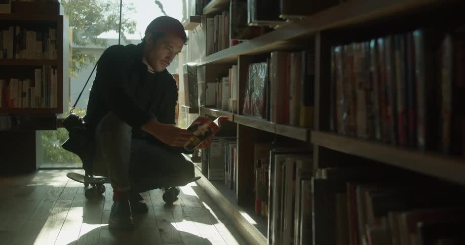 A man kneels on the ground as he flips through a book at the library