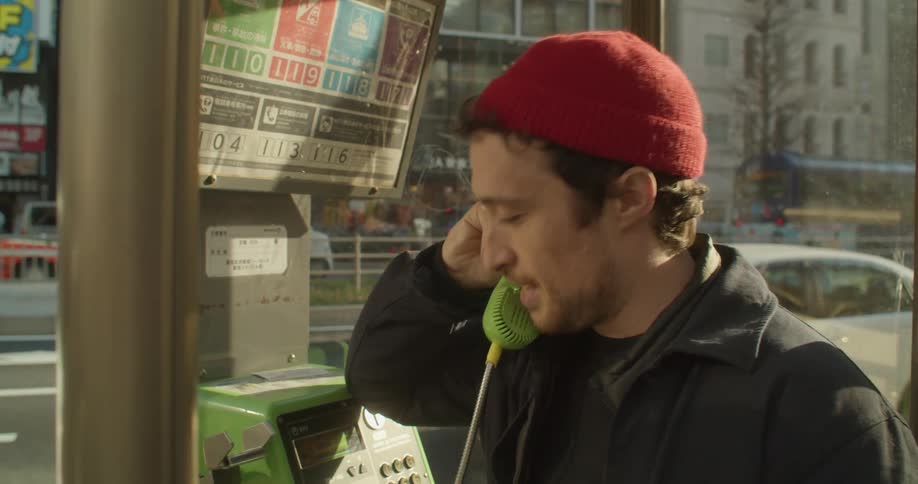 A man talks on a payphone as cars drive by on a city street on a sunny day