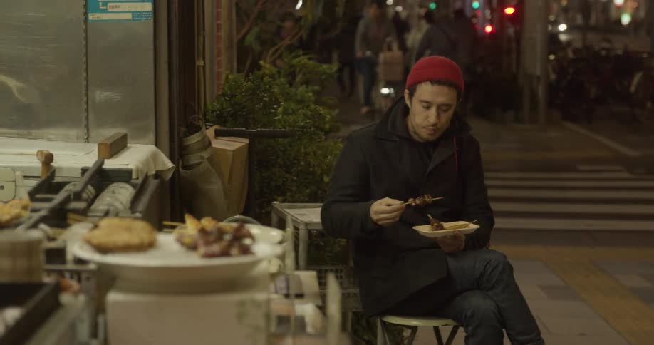 A white male sits on a stool and eats food on a busy city sidewalk
