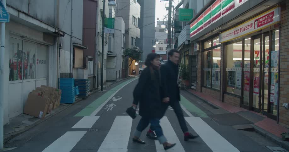 A white couple walk across a narrow street in a city