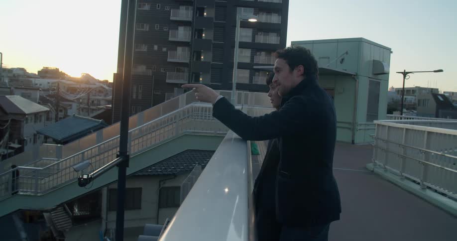 A man stands beside a woman on a platform above the city and points and talks