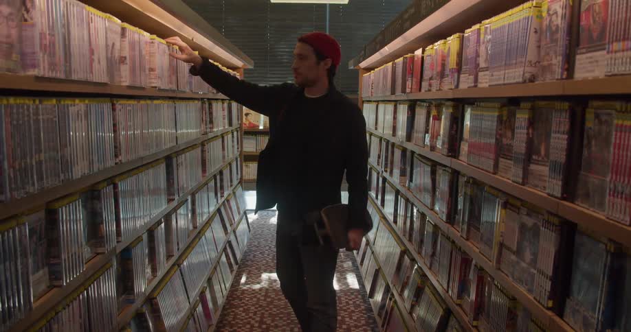 Male carrying a skateboard searches for a movie on the shelf in the video section of a store