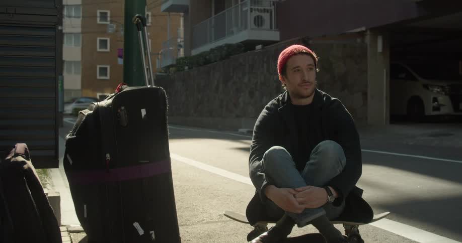 A man sits on his skateboard with luggage beside him on a neighborhood street