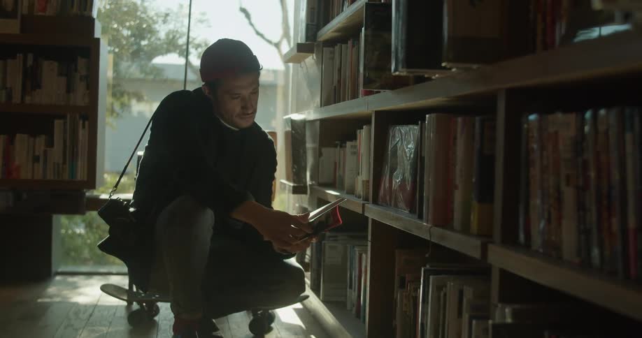 A man with a camera slung over his shoulder and a skateboard behind him is squatting down and flipping through a book in a book store