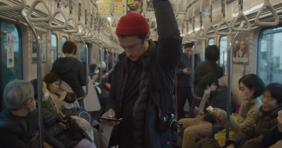 A man stands on a subway train holding onto a handle with one hand while scrolling on his phone with the other