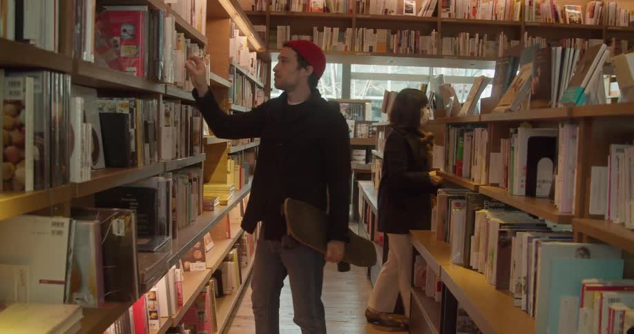 A white male walks through a library and looks at a book