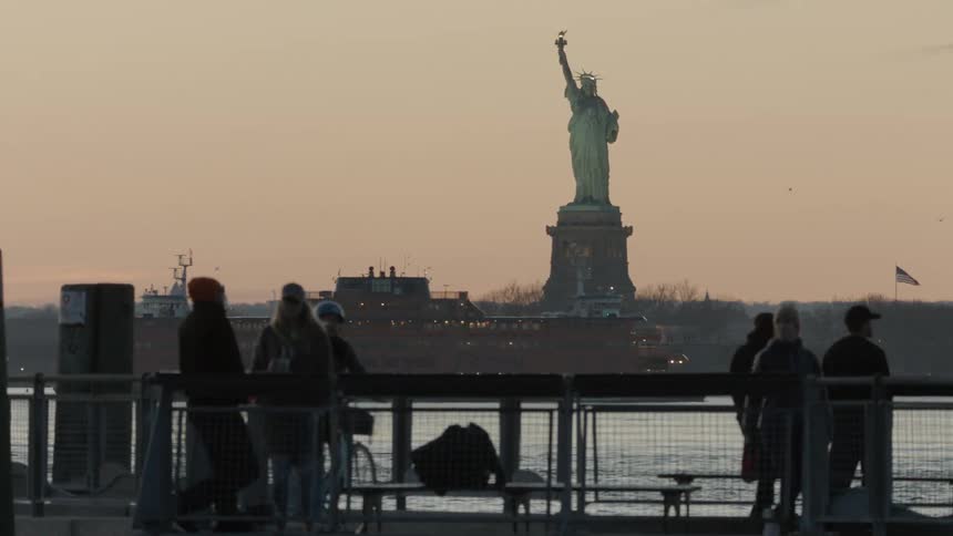 People walk along on the promenade while the ferry passes by the statue of liberty at dusk