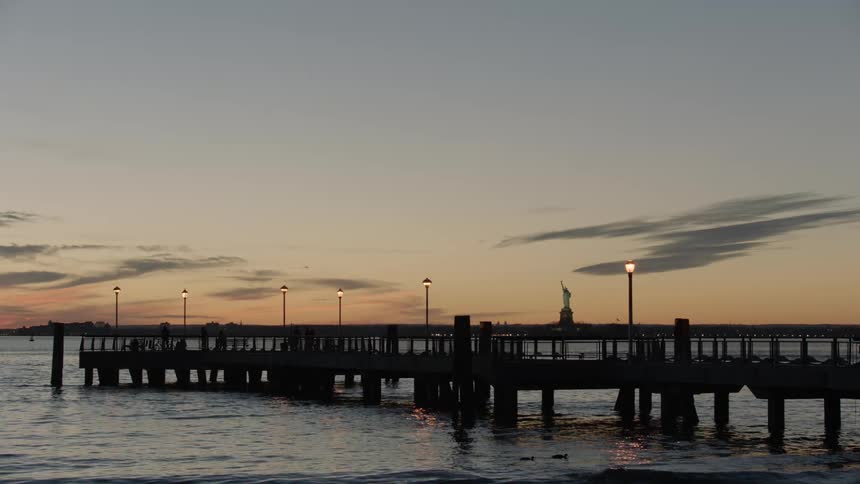 The statue of liberty sits in the distance behind a pier at sunset