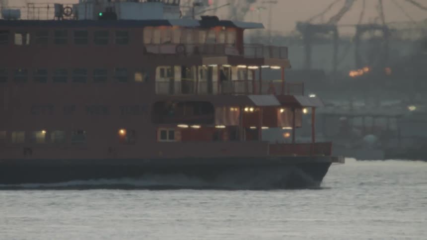 A three level ferry travels in the water past a shipping yard
