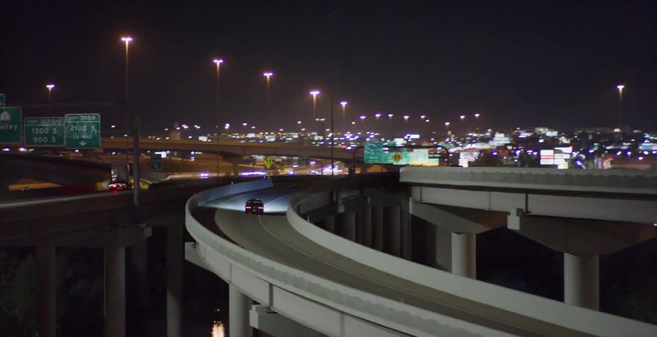 A car with its headlights on drives on an elevated roadway at night outside a city