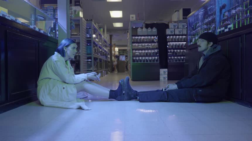 A man and woman sit in an aisle of a grocery store with their feet touching