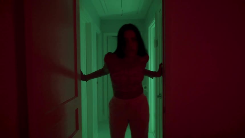 A woman takes exaggerated steps down a hallway as green and pink lights flash around her