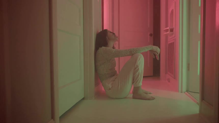 Barefoot woman sits on the floor of a hallway lit by colorful lights