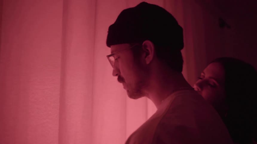 A woman puts her head on her boyfriends shoulder as they stand next to a large window in a room bathed in pink light