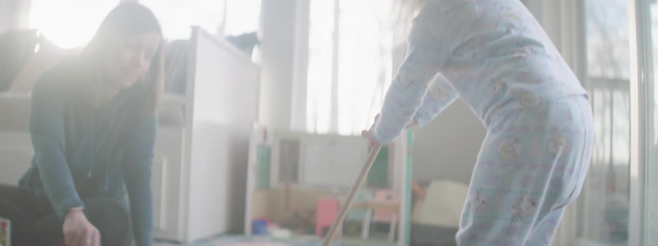 Small child sweeps with a toy broom and pushes the broom into a dustpan held by her mother