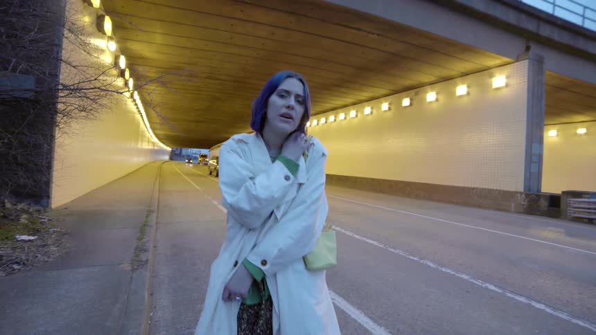 A white woman with an overcoat walks on the side of a road by a tunnel