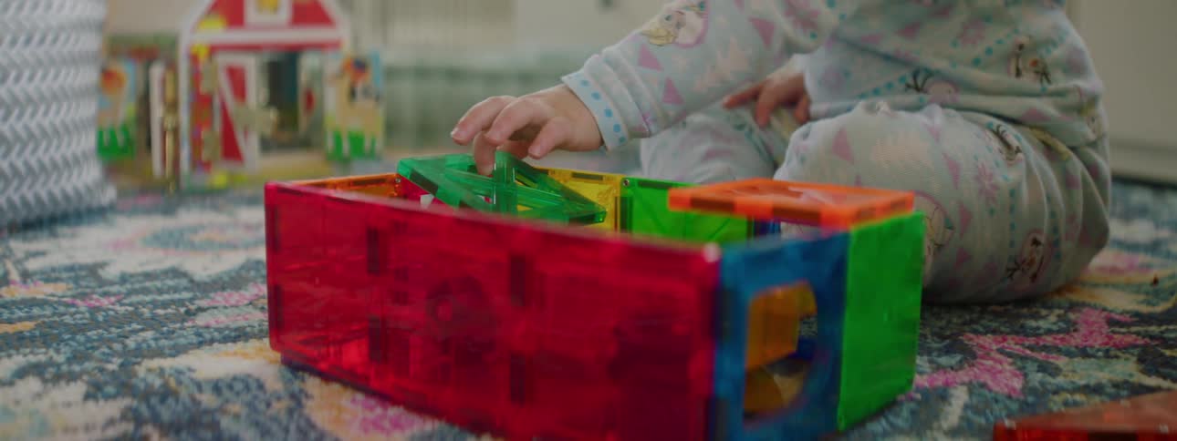 A little girl sits on the rug in her bedroom playing with her toys with a parent sitting cross legged beside her