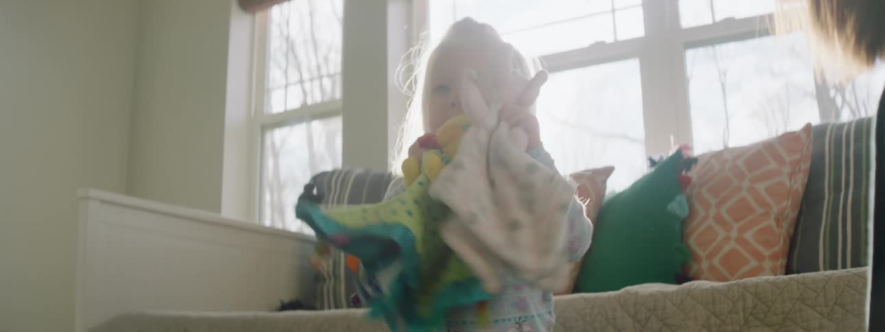A young child plays with toys in a living room while her mother looks on