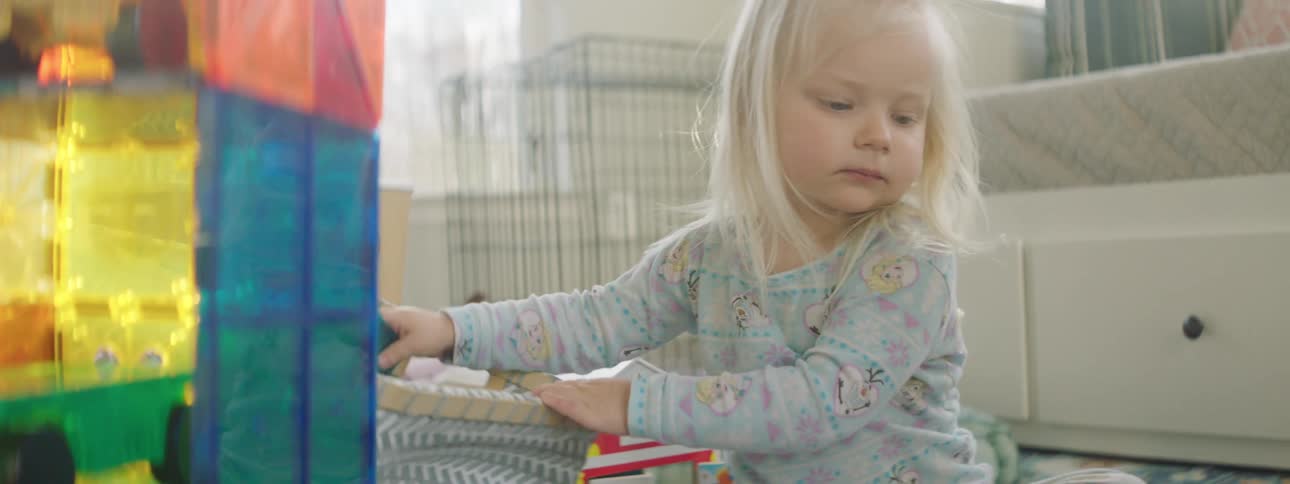 A toddler plays with toys in a bedroom with a parent helping