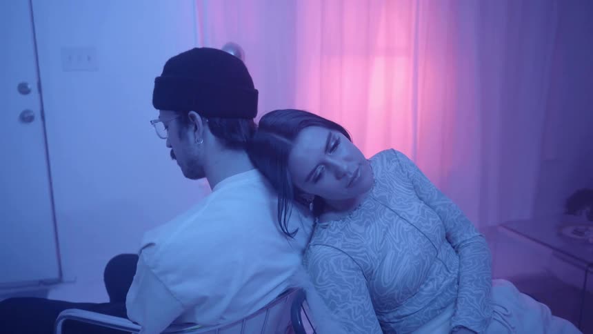 A woman sitting in a chair back to back with a man opens her eyes and looks up while a pink light glows in the window