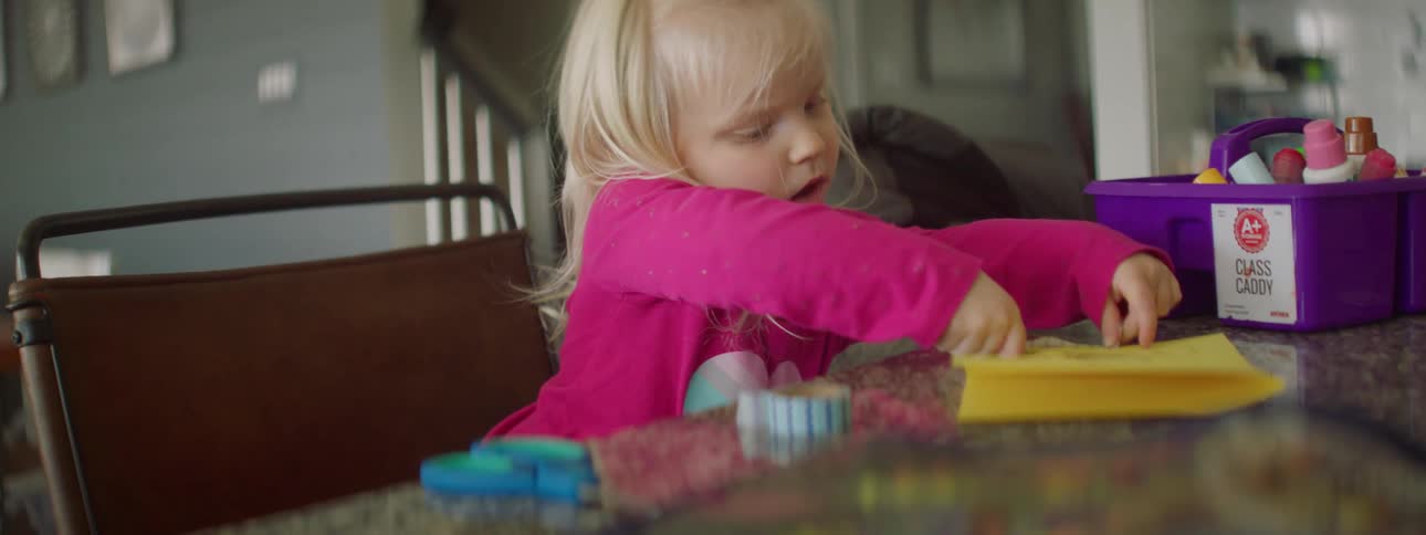 A little girl sits at a table playing with a sticker and a piece of paper