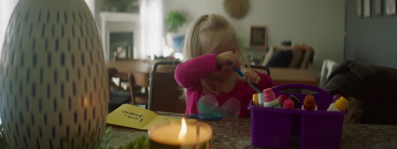 Young blonde child removes something from a basket as she sits at a table with a burning candle
