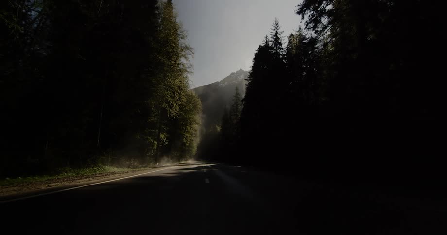 The sun shines and glares over the mountain peak and through the trees on an empty rural road
