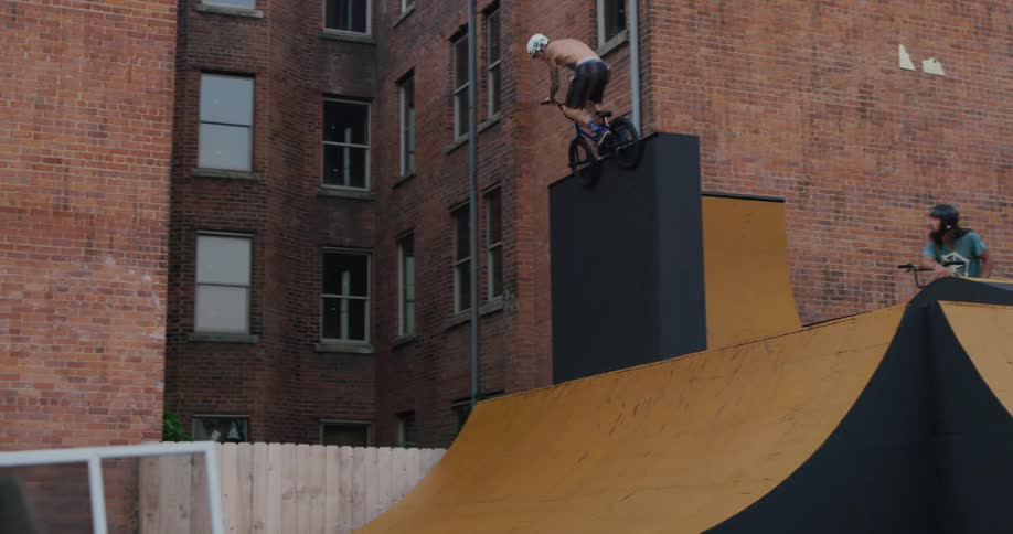 Two friends watch as another makes a jump and holds his bike still on a ramp behind an apartment building
