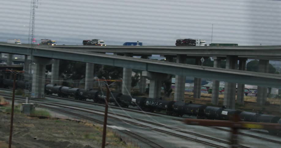 A train below a road with large trucks and cars driving