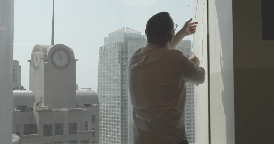 A man raises the blind in a large office window showing the city and clock tower outside