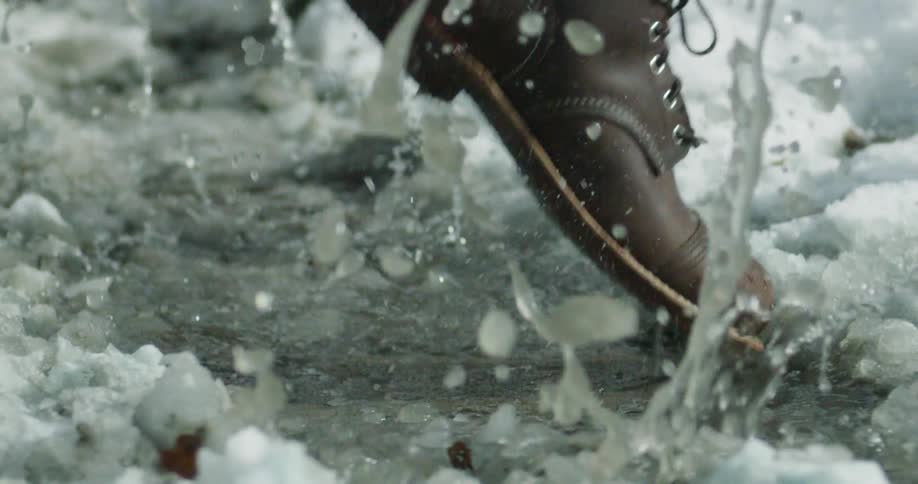 A booted foot steps into a small stream of water, ice and snow