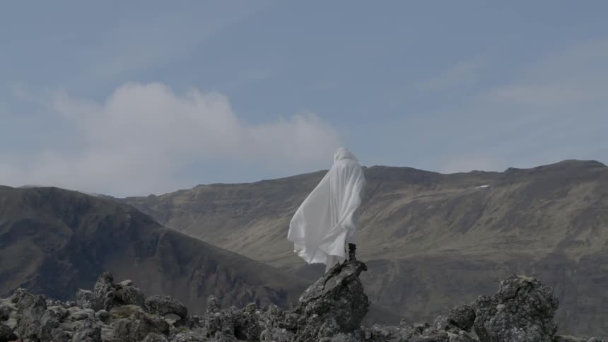 A figure all in white standing atop a rock pile with a range of mountains in the background