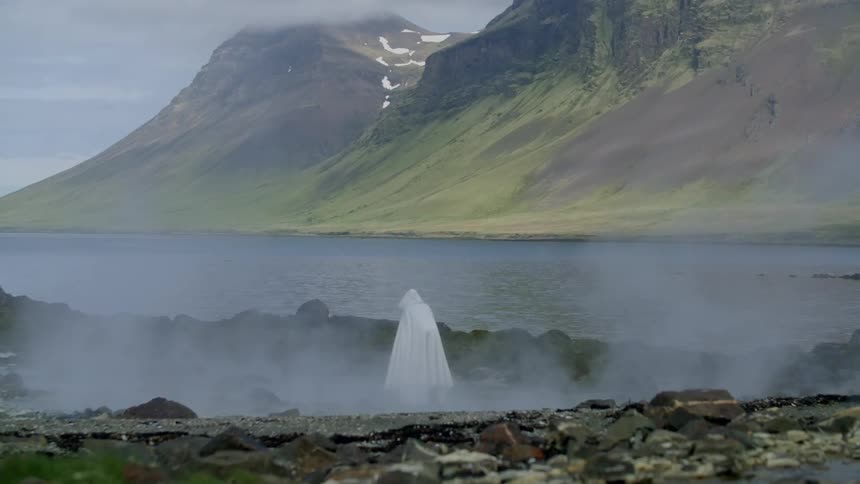 A cloaked figure stands near a mountain lake