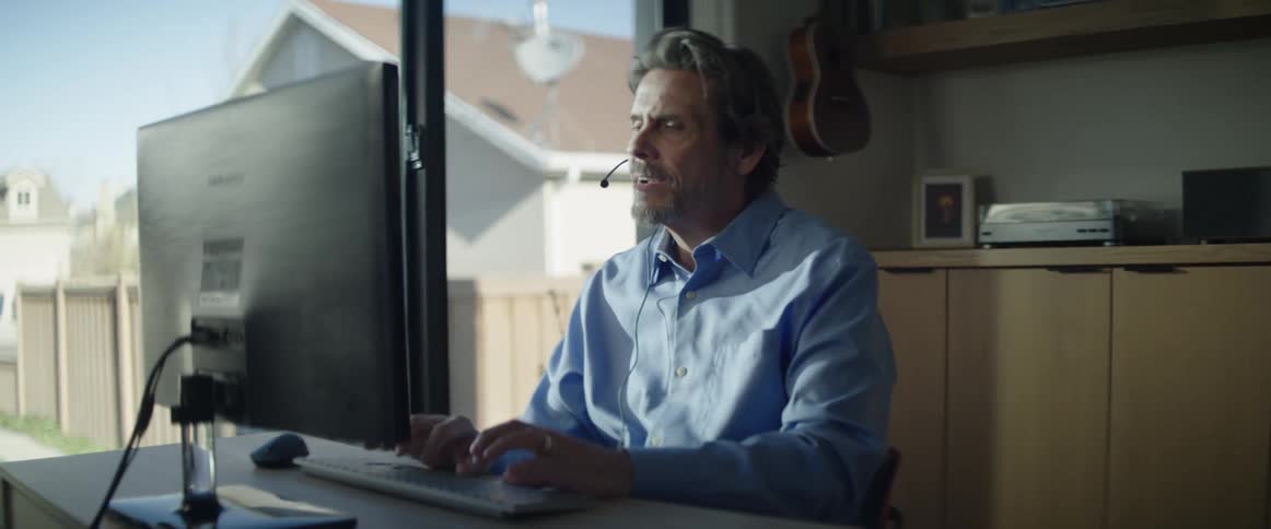 A man sitting at a desk using a computer while talking into a headset