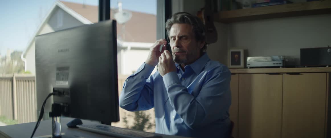 Male adjusts headset with microphone as he is talking and looking at the computer monitor in his home office