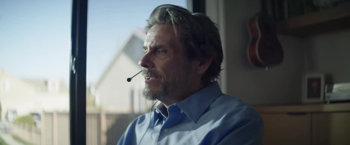 An older white man sits in his home on a computer and talks to someone through a headset