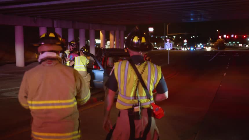 A pair of firefighters walking towards a vehicle where another group of firefighters are lifting a child from a car under a city bridge at night