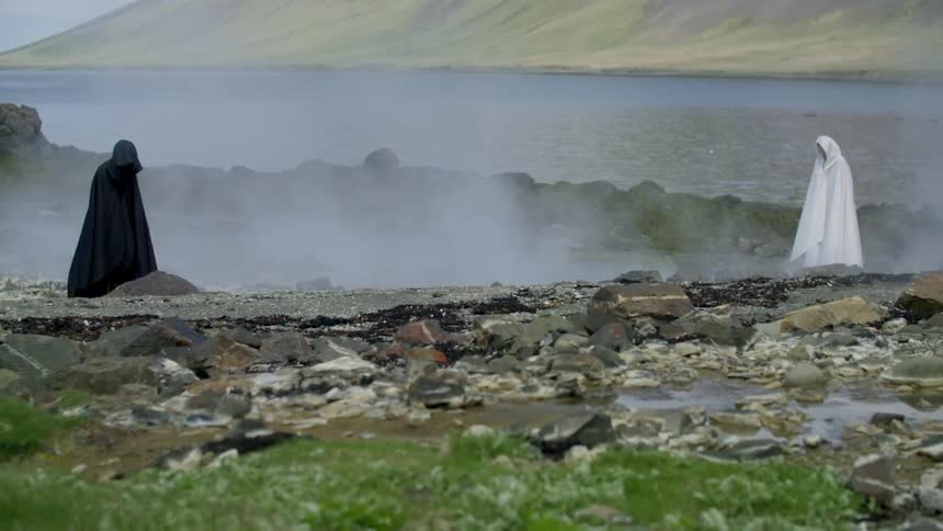 A black caped figure stands along a rocky ledge in the fog