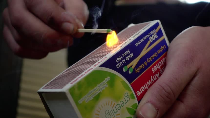 A white man strikes a wooden match on a strip on a box