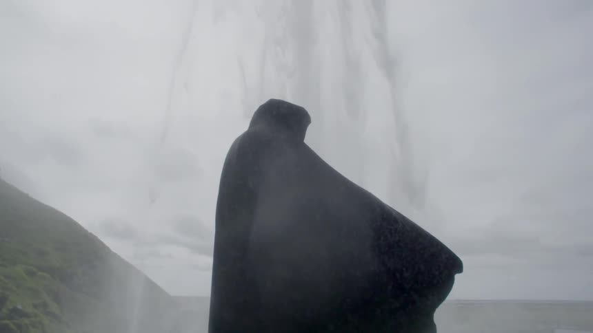 A person wearing dark robes is standing below a waterfall on a cloudy day