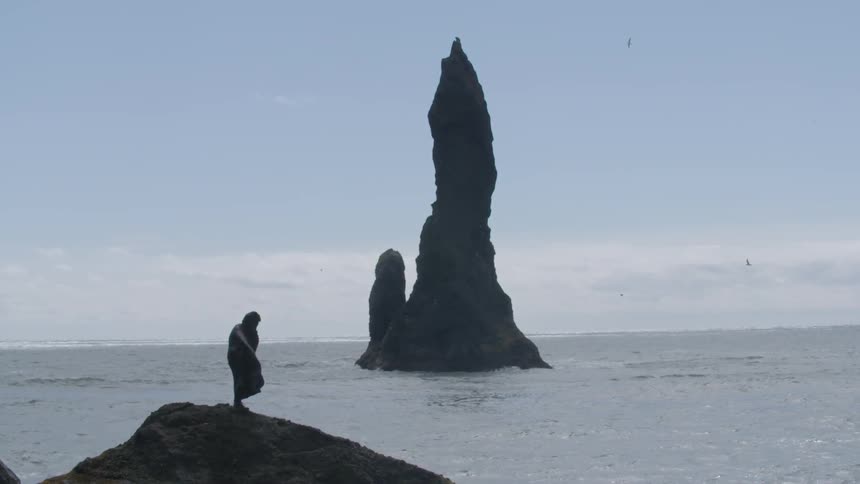 A cloaked figure stands on a rock overlooking the ocean with a large rock formation in the distance
